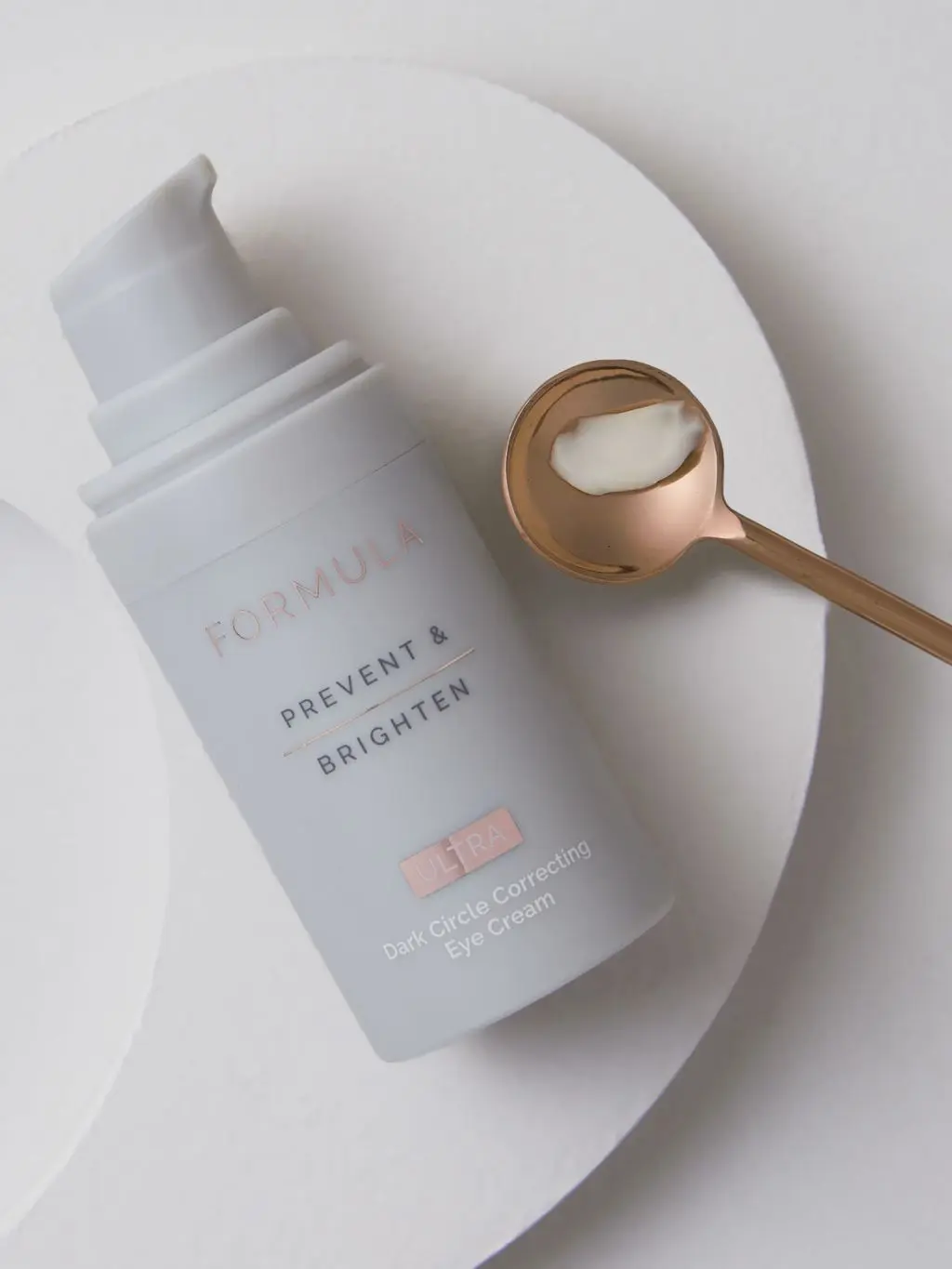 A bottle of Formula Brightening eye cream lies on a plate next to a copper spoon containing some of the eye cream