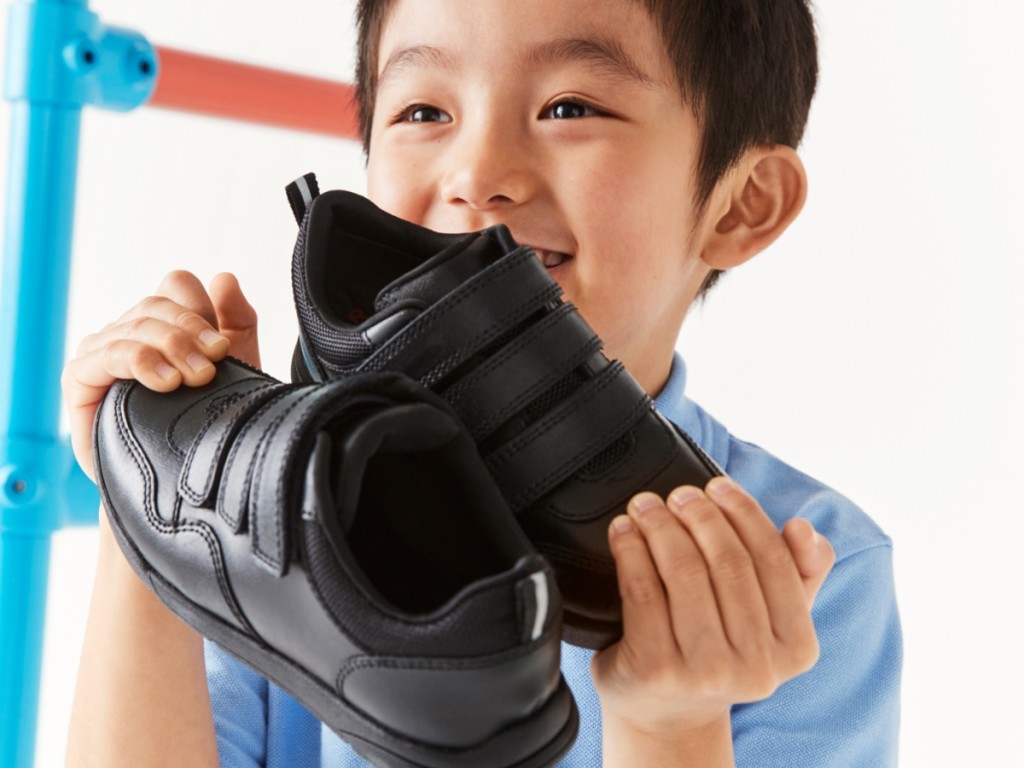 Boy wearing a blue short-sleeved shirt holding black school shoes. Shop school shoes