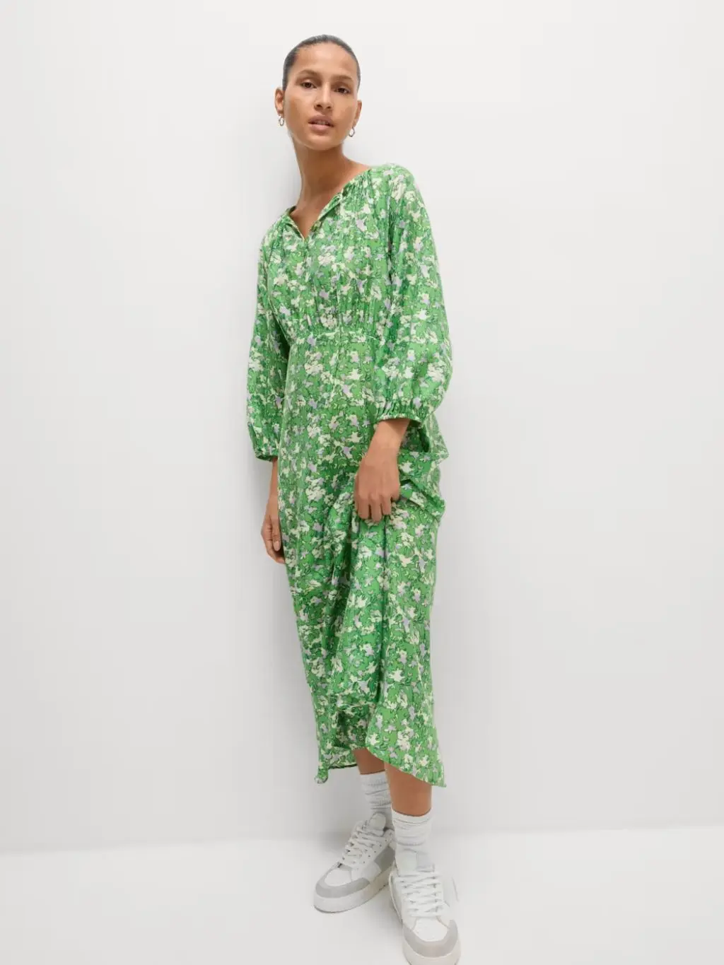 A woman wears a green floral dress with white trainers