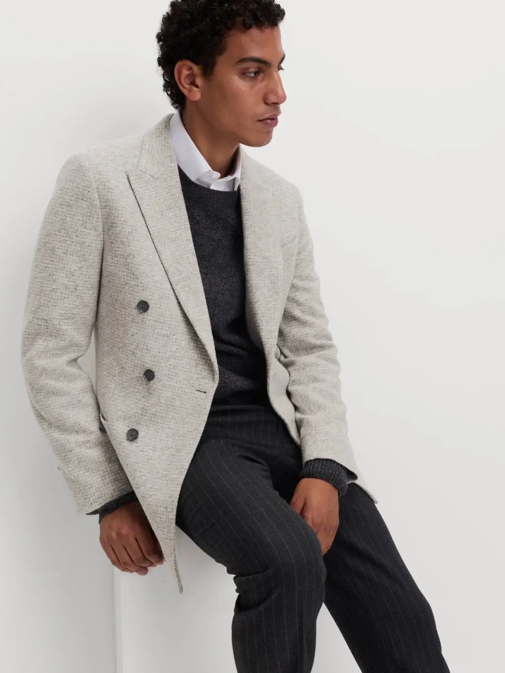 A man wears a pale wool blazer over a white shirt and crew neck jumper, matching it with pinstripe trousers. He's sat on a stool against a pale background