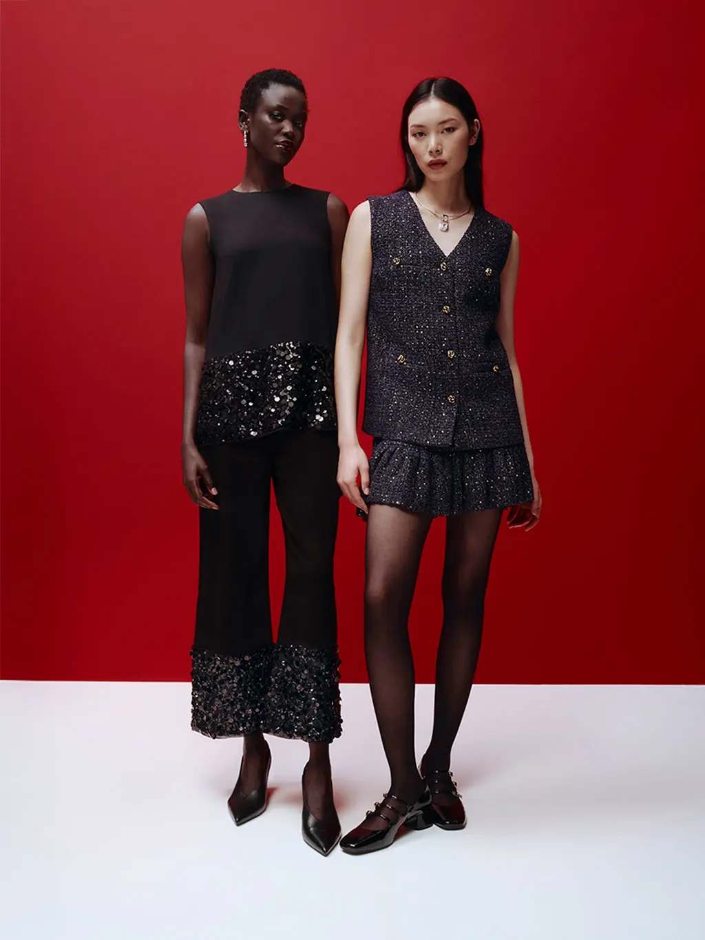 Woman wearing a sequinned top and trousers with heels, beside a woman wearing a glittery waistcoat and skirt with heels.