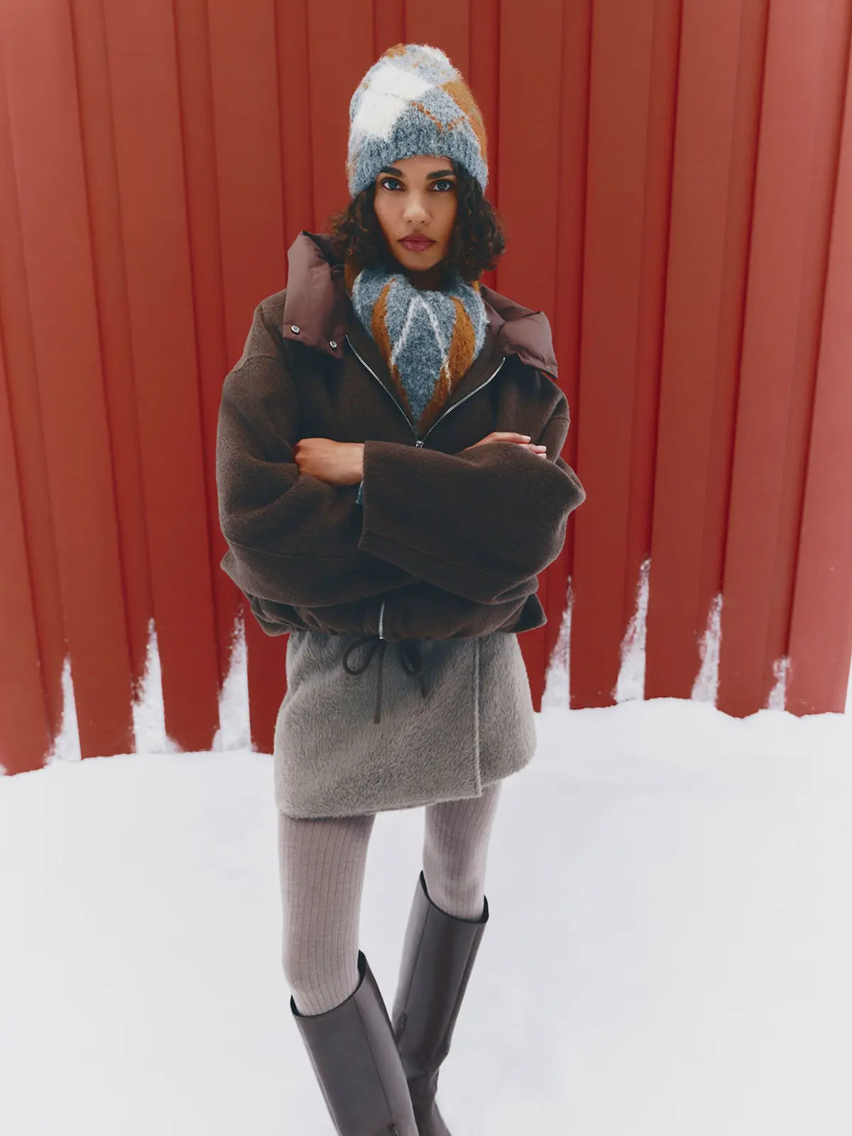 Woman wearing brown jacket, grey mini skirt, tights, tall boots, argyle-patterned hat and matching scarf