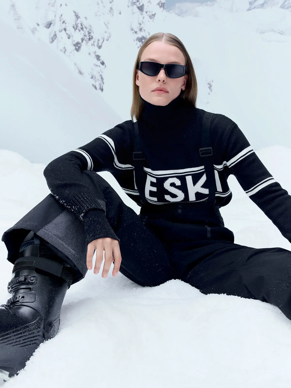 Woman wearing black jumper with white slogan, black ski trousers, ski boots and black ski glasses
