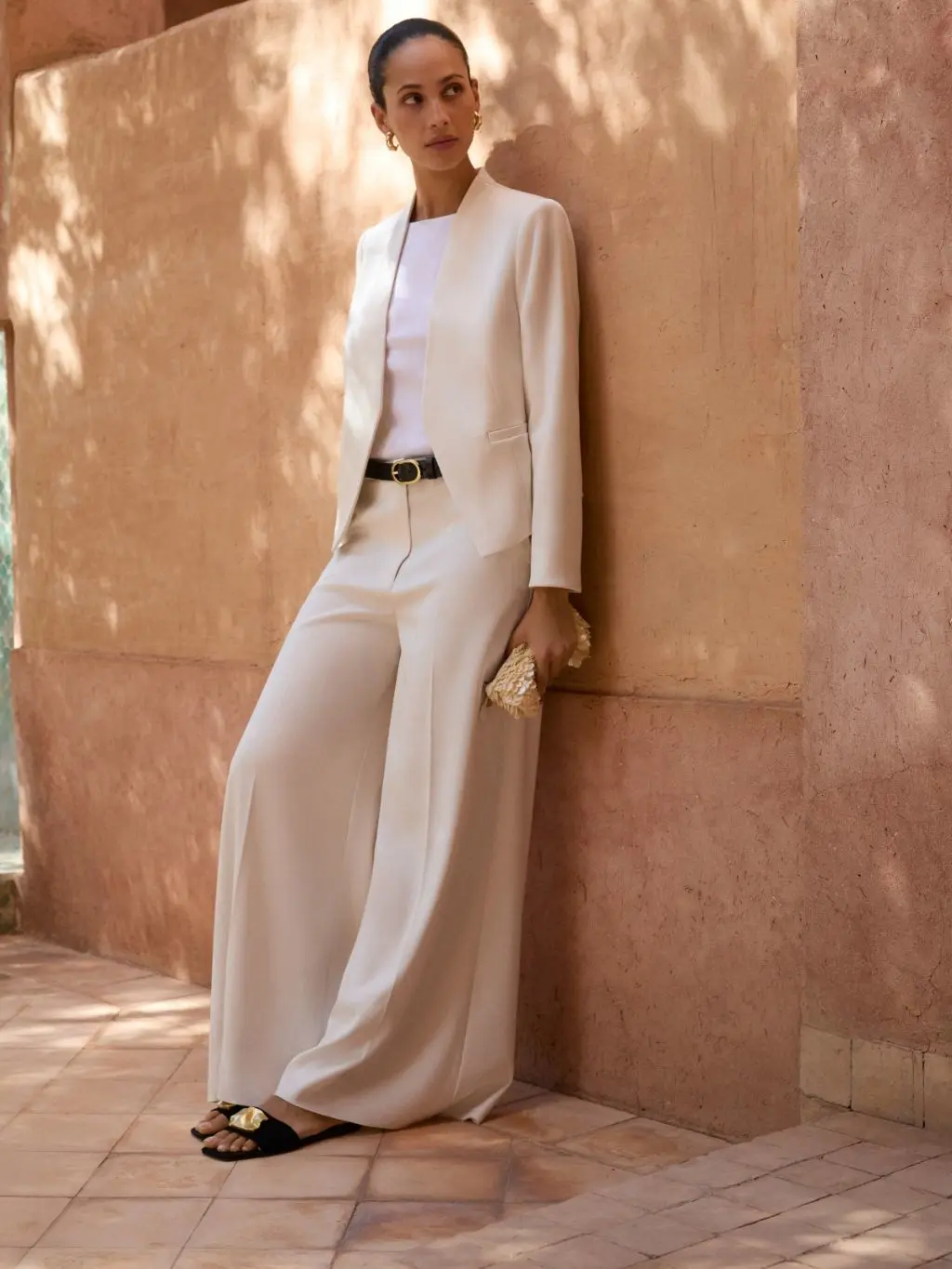 A woman leans against a wall wearing a cream wide leg trousers suit and white t-shirt