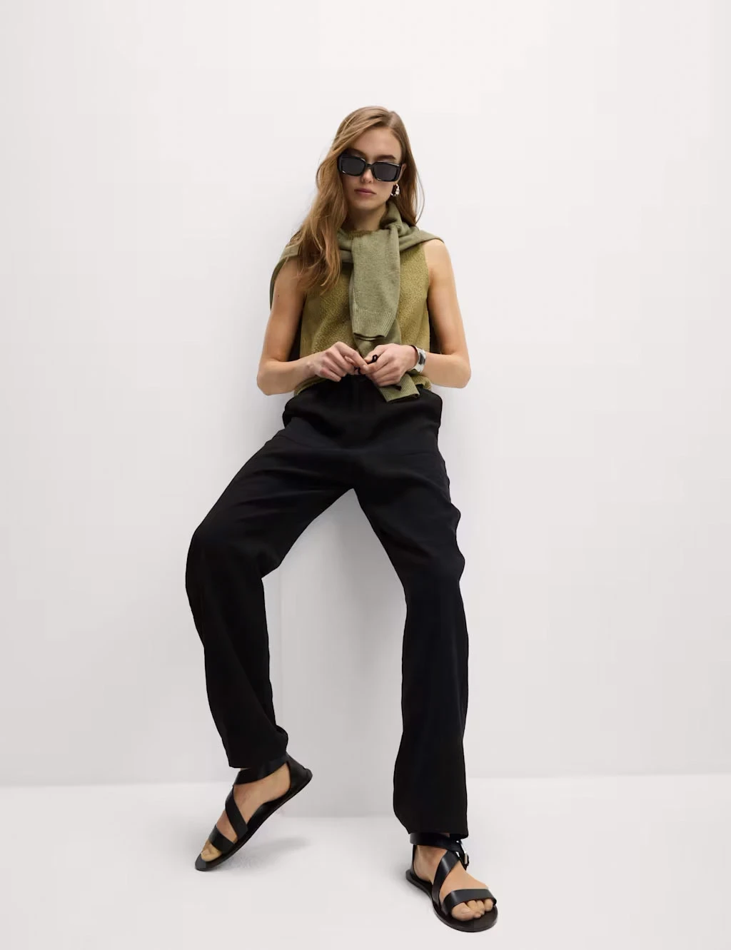 Woman wearing a green vest with black trousers, black sandals, sunglasses and a green jumper over her shoulders.