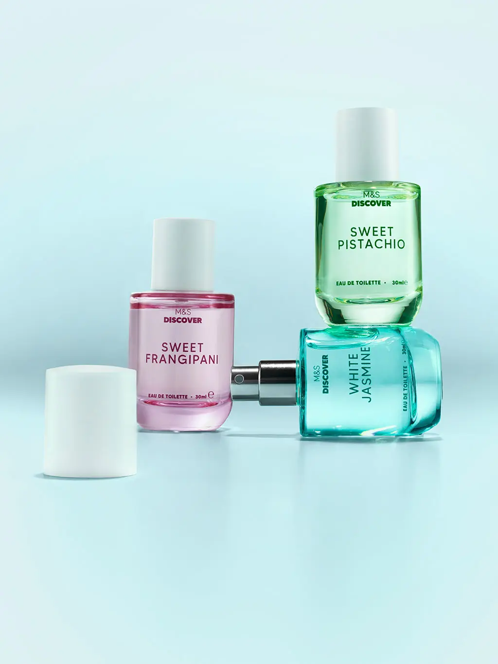 Group of three M&S perfumes in pink, green and blue glass bottles on a blue background.
