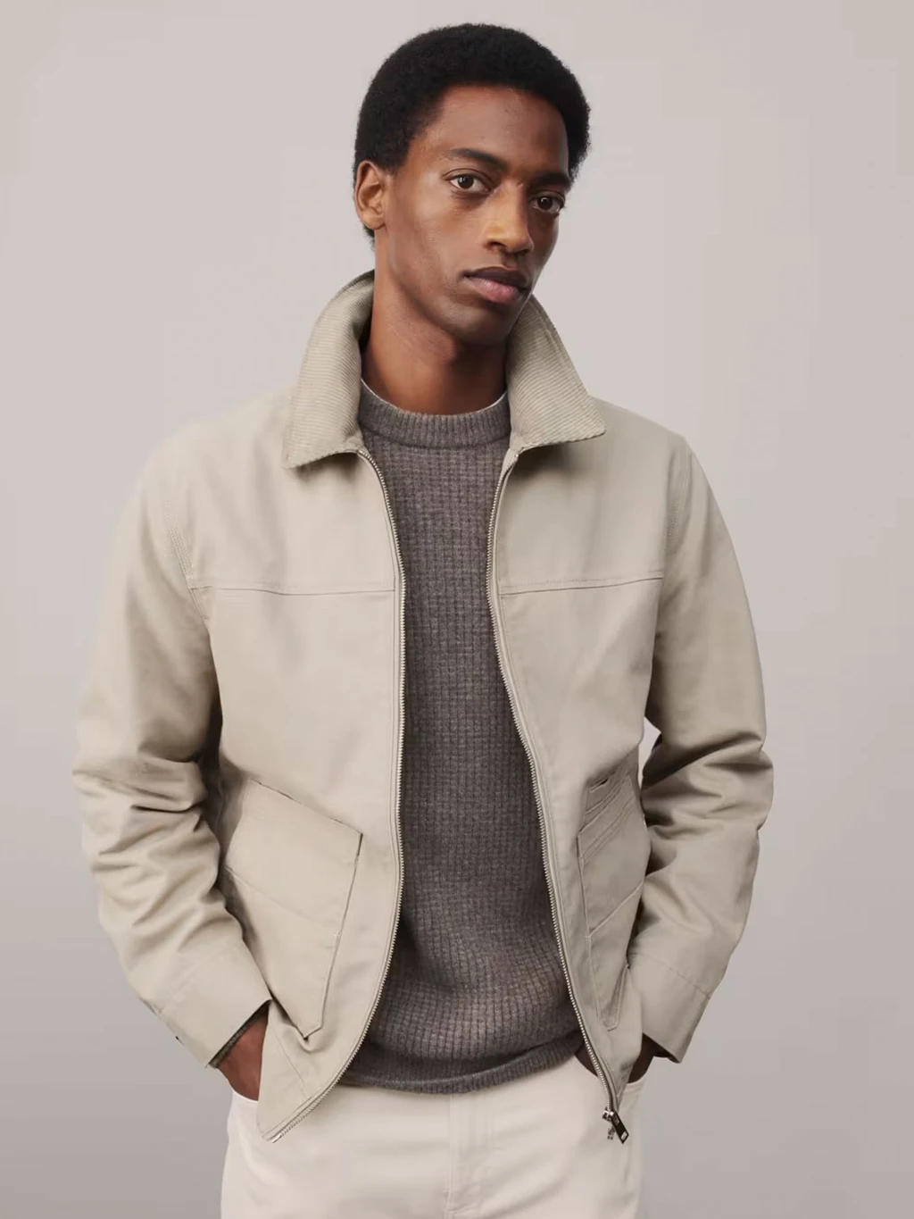 A man wears a cream utility jacket with a jumper and ecru trousers