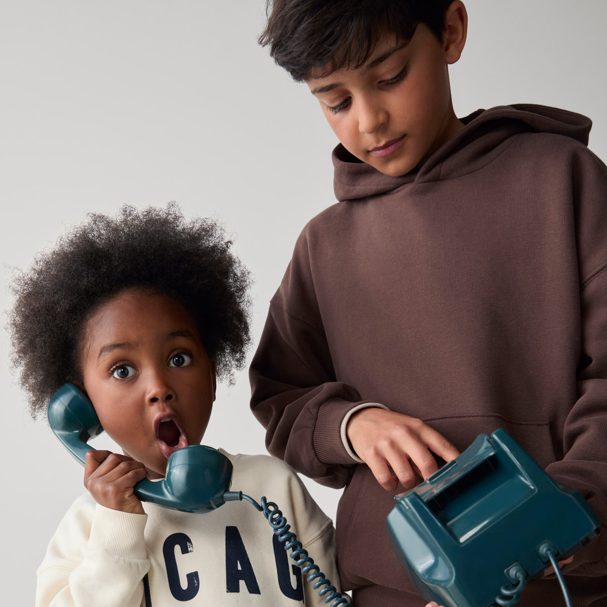 Two boys wearing hoodies playing with a teal vintage telephone