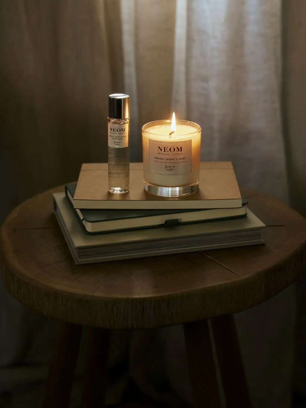 A bedside table with books, skincare and a lit Neom candle on it