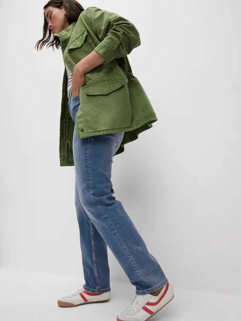 A woman wears a khaki jacket, straight leg M&S jeans and red and white trainers