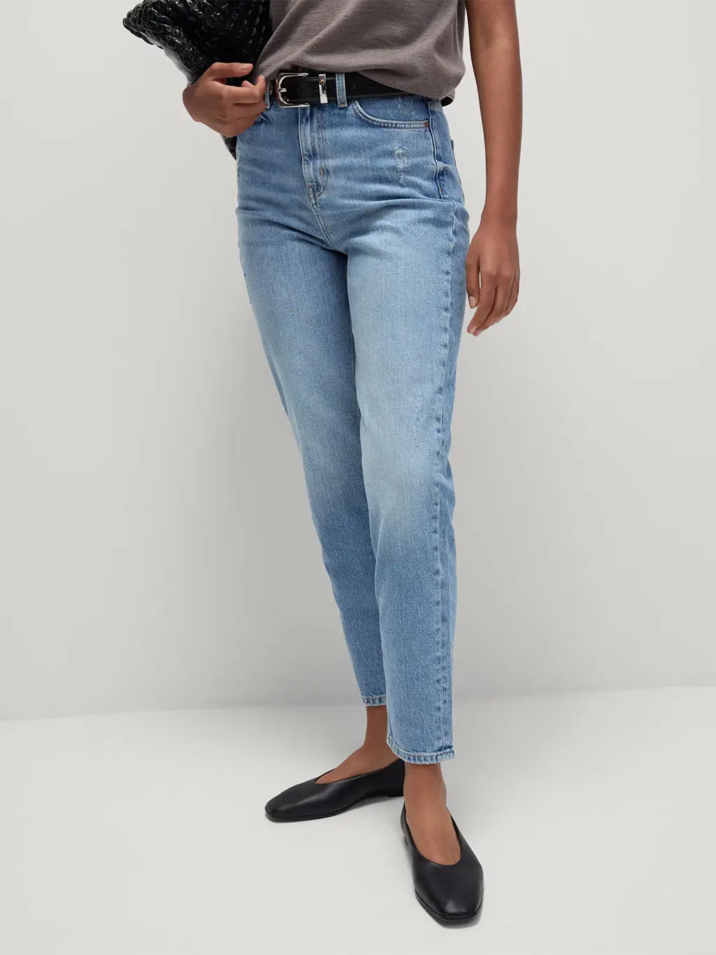 Woman wearing light blue mom jeans with a taupe vest, black belt, black ballet pumps and a black tote bag