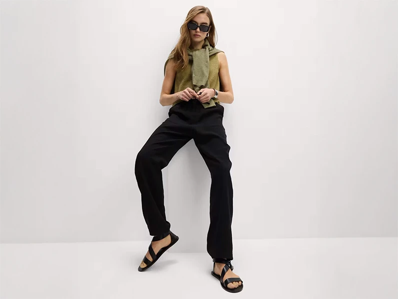 Woman wearing a green vest with black trousers, black sandals, sunglasses and a green jumper over her shoulders.
