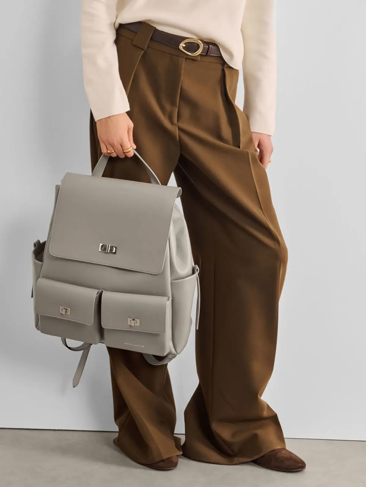 Woman wearing smart brown trousers and neutral knit, holding a grey backpack
