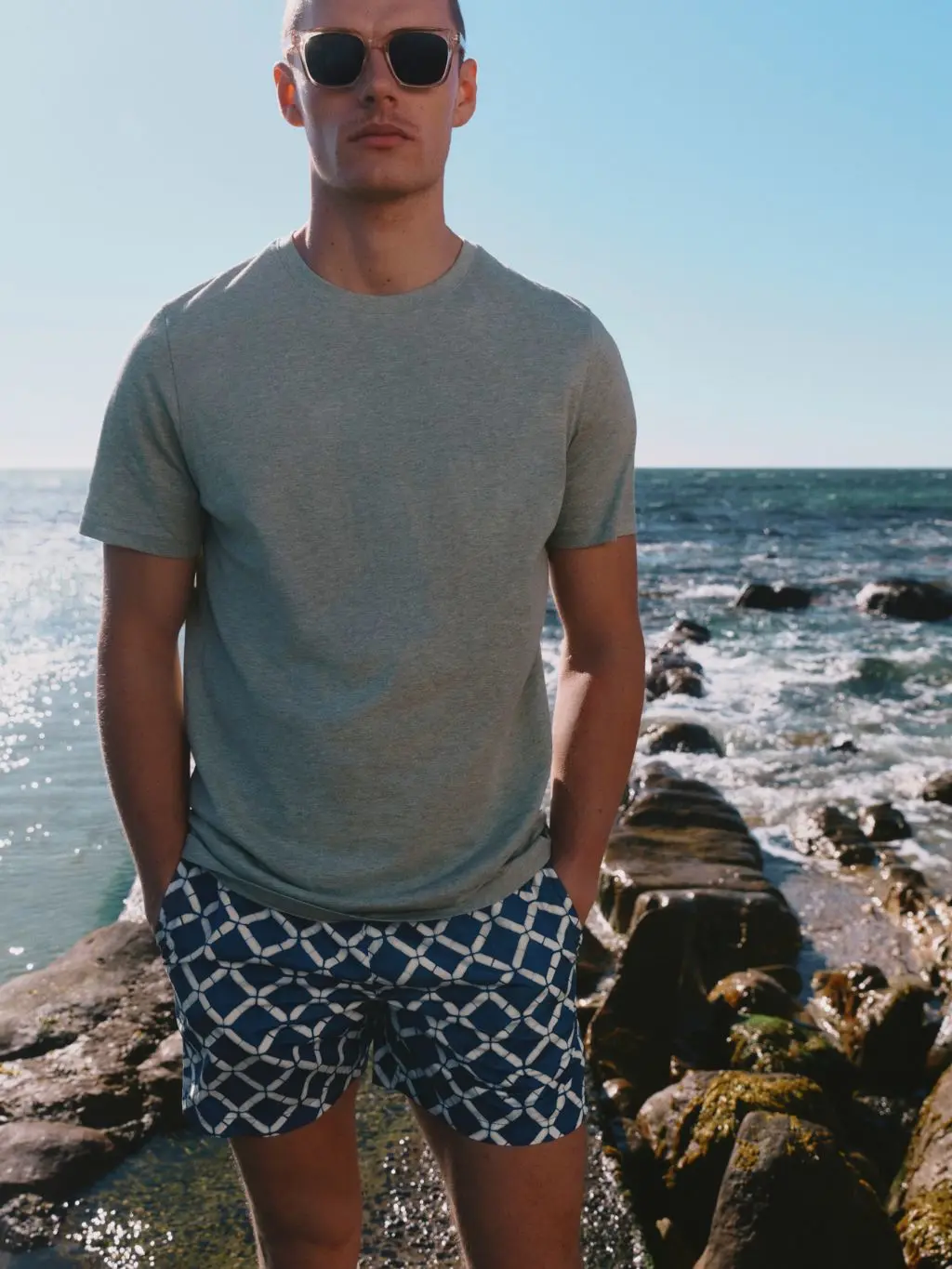 A man wears a t-shirt and swimming shorts while stood against a backdrop of the sea and some rocks