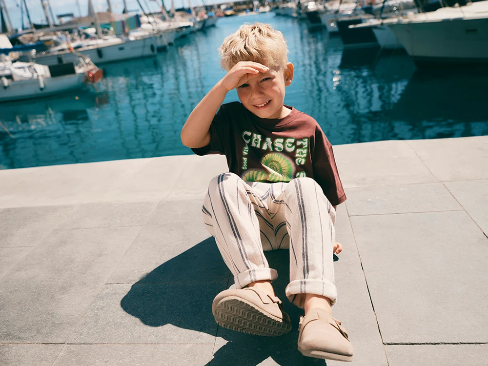 A boy wears a brown T-shirt, cream striped trousers and beige clogs