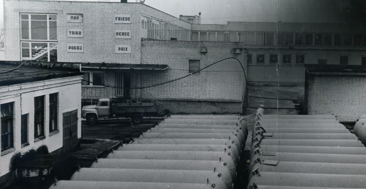 Archyvinė sulčių gamyklos nuotrauka. 1982 metai. Matomos cisternos ir įvairiomis kalbomis ant sienos parašytas žodis "taika".