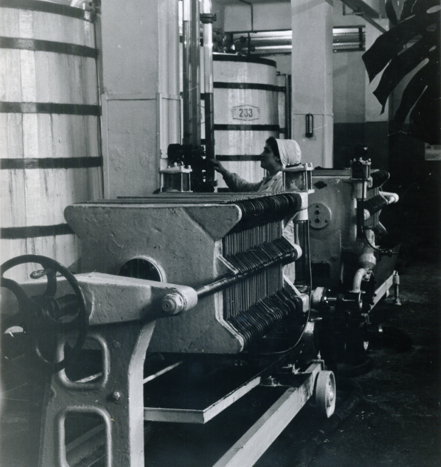 Archyvinė nuotrauka, sulčių spaudykla iš 1984 m. Matosi didelės statinės, prie kurių moteris dirba su įranga.