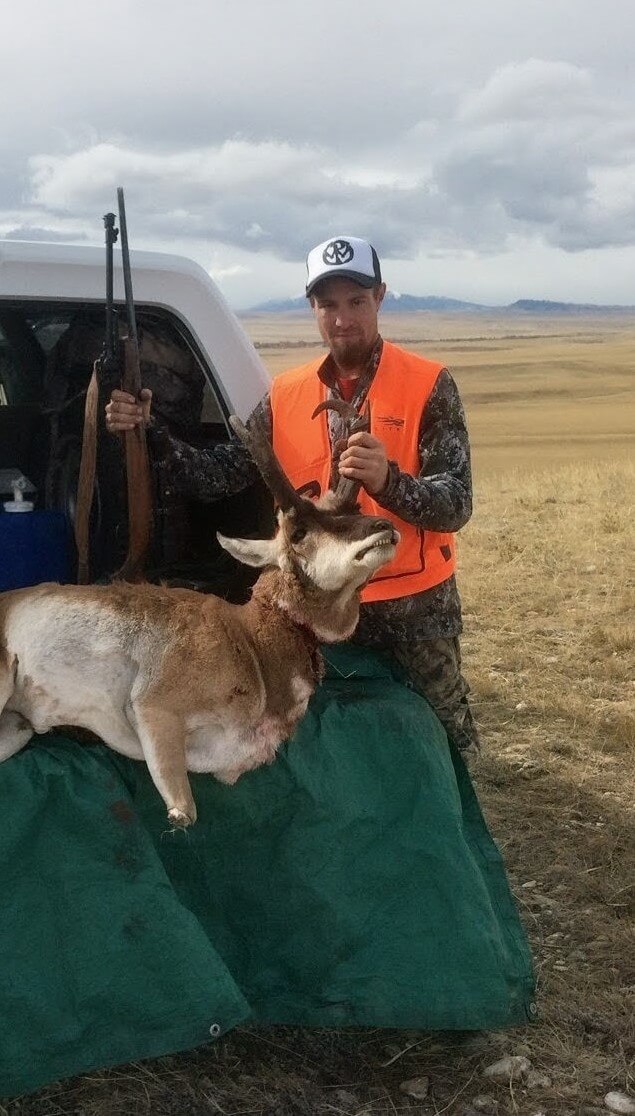 Hunter in orange vest holding pronghorn antelope and rifle by pickup tailgate