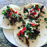 Three shredded meat tacos with crumbled cheese, tomato slices and chopped cilantro on a white plate