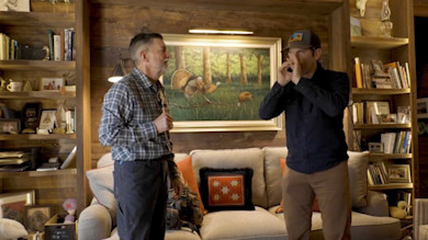 Man demonstrating a crow call to another man in a wood-paneled room with sofa and turkey painting
