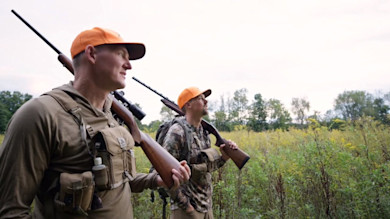 Two hunters in orange caps carrying rifles over their shoulders in a grassy field