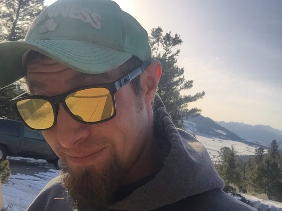 Man in green 'GUINNESS' cap and yellow sunglasses, snowy mountains behind him