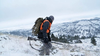 Hunter in orange vest carrying rifle and large backpack across snowy badlands ridge