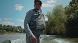 Man standing on boat wearing gray NRS life vest, cap and sunglasses on river