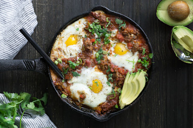 Cast-iron skillet with chorizo shakshuka, three baked eggs, sliced avocado and cilantro
