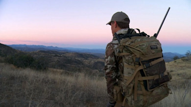Hunter with rifle and camo pack overlooking rolling hills at pink dawn
