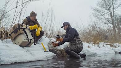 Beaver trap set by two men at snowy creek, one kneeling in the water
