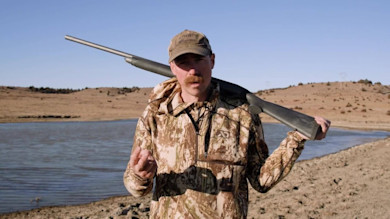 Hunter in camouflage jacket and cap with shotgun slung over shoulder by pond
