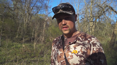 Hunter wearing First Lite camo shirt and cap, sunglasses on cap, standing in leafless woods
