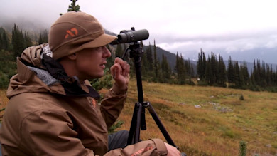 Steven Rinella looking through a spotting scope on tripod in alpine meadow, First Lite hat