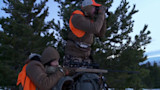 Two hunters in orange vests at dusk—one crouched aiming a rifle from a pack, the other standing with binoculars