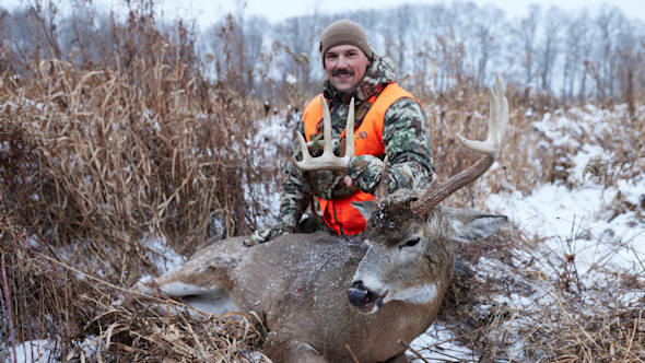Hunter in camo and orange vest kneeling beside large buck in snowy field, holding antlers