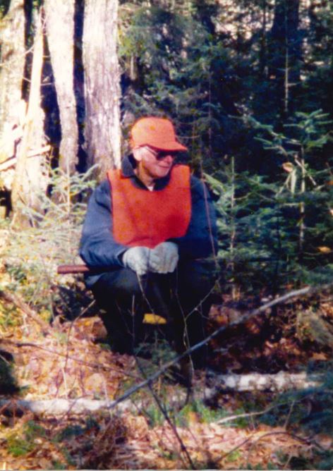 Man squatting in forest wearing orange cap and safety vest, holding a rifle