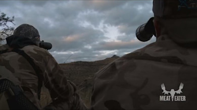 Two hunters looking through binoculars toward a distant ridgeline under cloudy sky, MEAT EATER logo
