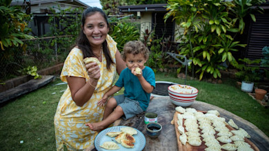 Woman and toddler making and eating fish gyoza outdoors, wooden table with rows of folded dumplings