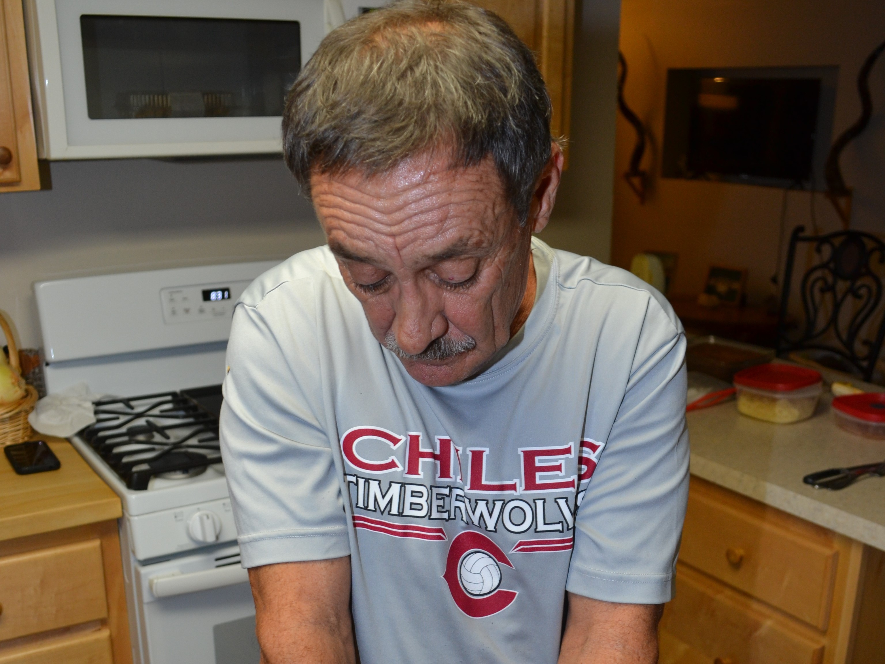 Older man leaning over kitchen counter wearing shirt reading 'CHILES TIMBERWOLVES'