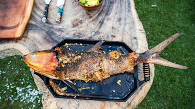 Whole stuffed fish on a black roasting pan atop a wooden slab, knives and avocado bowl nearby