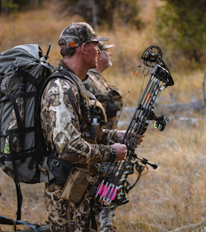 Two hunters in camouflage; foreground hunter holds compound bow with pink-fletched arrows and a large backpack