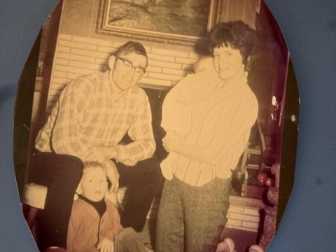 Family portrait: seated man in glasses, woman holding infant, young child on floor by fireplace.