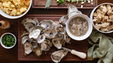 Shucked oysters on wooden board with bowl of oyster meat, bread, chopped herbs and a shucking knife