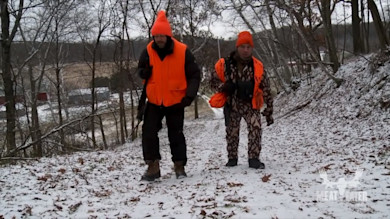 Two men in blaze-orange hunting gear carrying rifles walk a snowy farm road; MEAT EATER logo