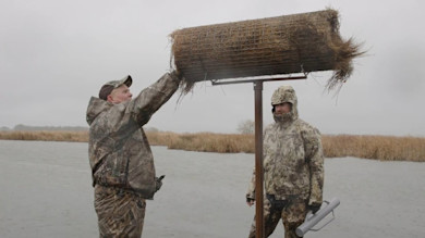 Two hunters placing a hay bale into a pole-mounted hen-house basket over a marsh