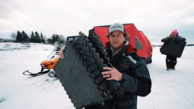 Man holding black foam pads on frozen lake by red ice shelter; hat reads CATCHandCOOK