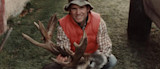 Hunter kneeling in orange vest and bucket hat, holding a deer with large antlers on grass