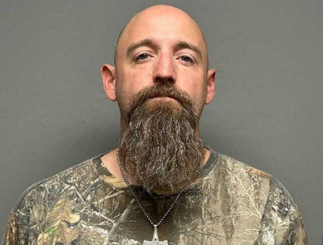 Bald man with long brown beard wearing camouflage shirt against gray background