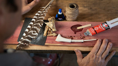 Hands assembling turkey wingbone call with feather, bone, twine, and J‑B Weld ClearWeld glue